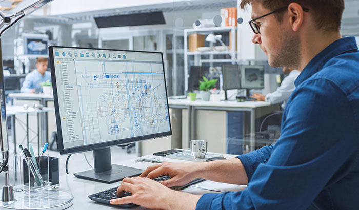 Over the Shoulder Shot of Engineer Working with CAD Software on Desktop Computer, Screen Shows Technical Drafts and Drawings. In the Background Engineering Facility Specialising on Industrial Design