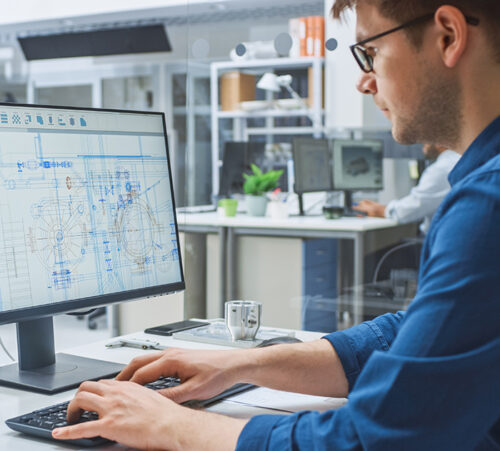 Over the Shoulder Shot of Engineer Working with CAD Software on Desktop Computer, Screen Shows Technical Drafts and Drawings. In the Background Engineering Facility Specialising on Industrial Design